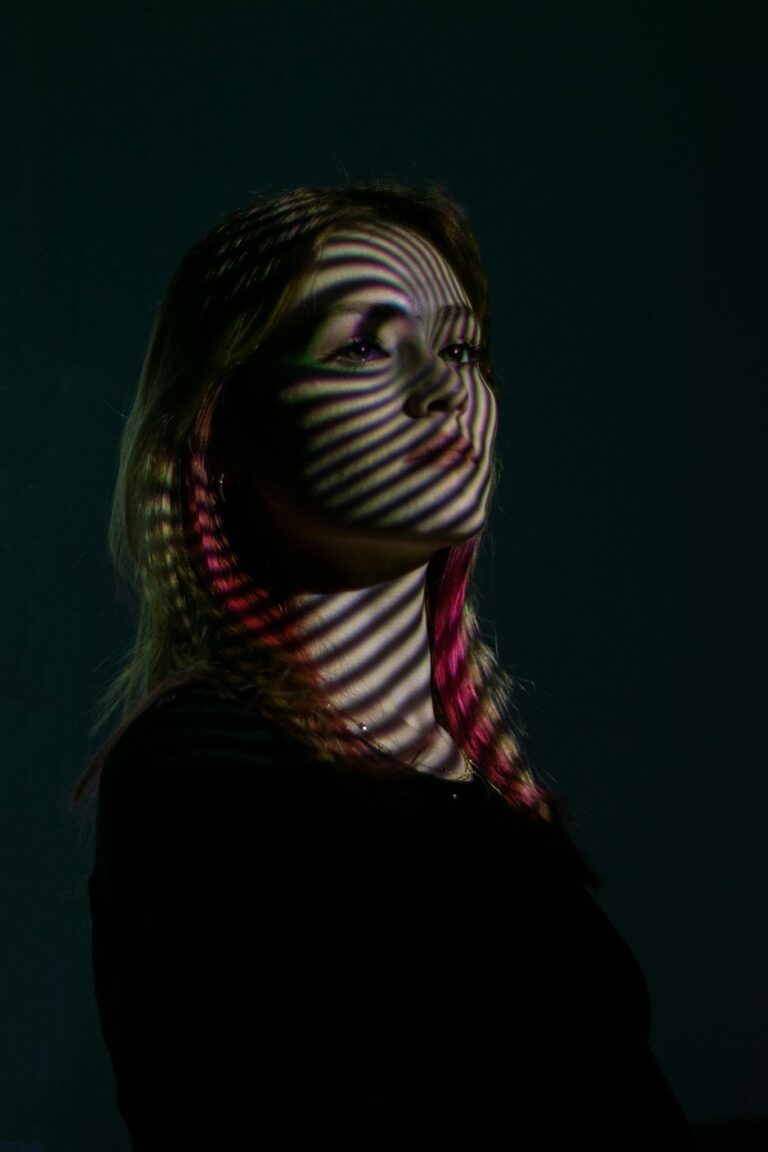 A woman in a dark setting with stripes of dramatic light creating an artistic shadow on her face.
