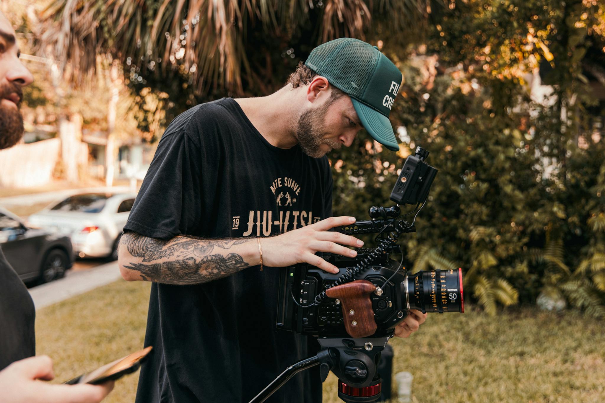 A young male cinematographer adjusts a cinema camera outdoors in a sunny setting.