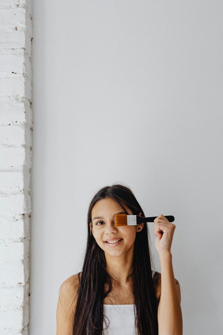 A young woman smiles while playfully holding a paintbrush indoors against a white background.