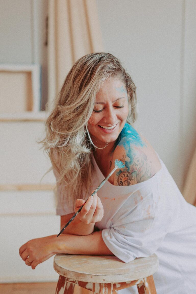 An adult woman smiling while applying blue paint on her shoulder with a brush.