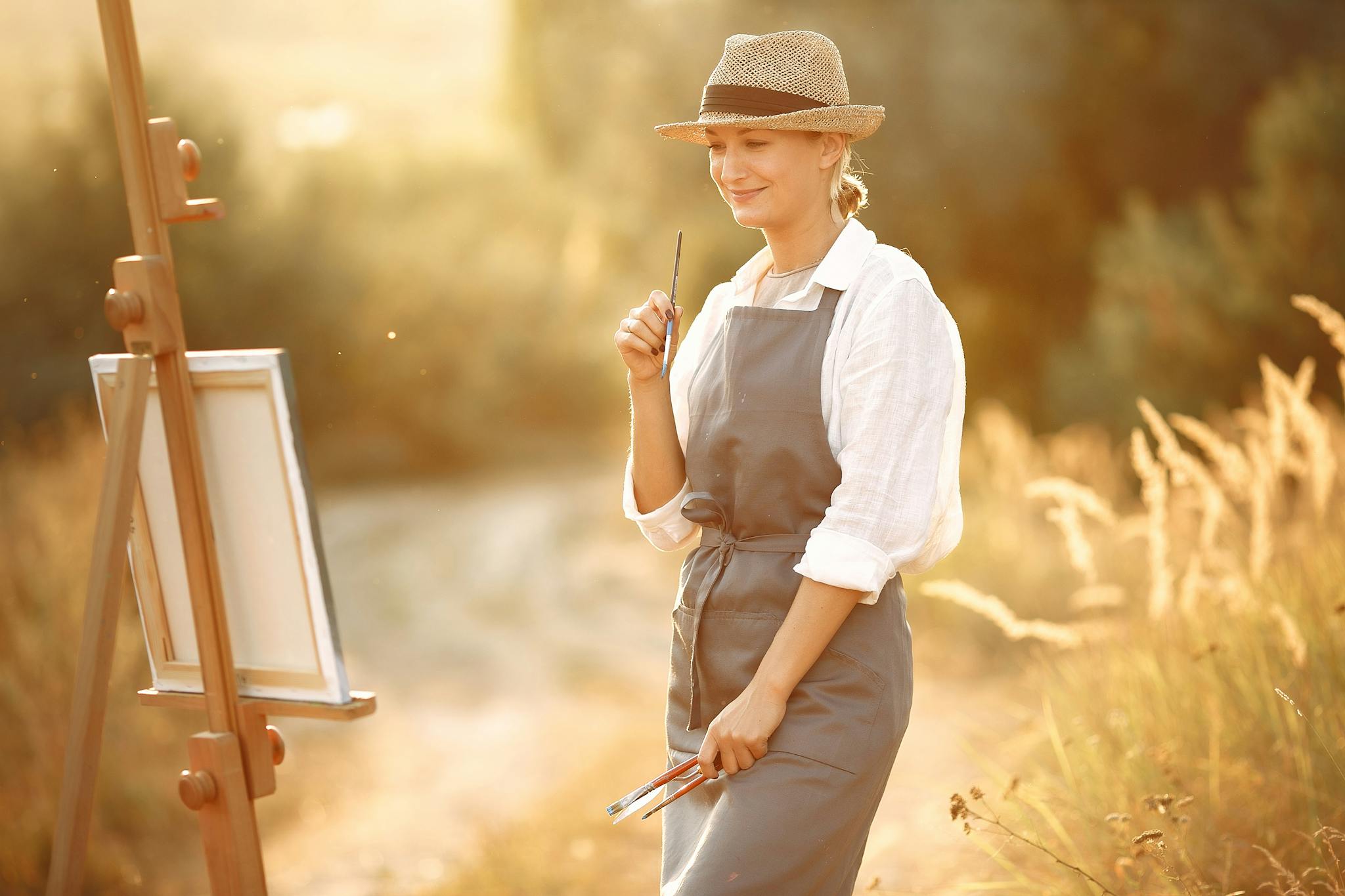 Side view of cheerful woman in hat and apron smiling and looking at own painting while standing in summer sunny day in nature