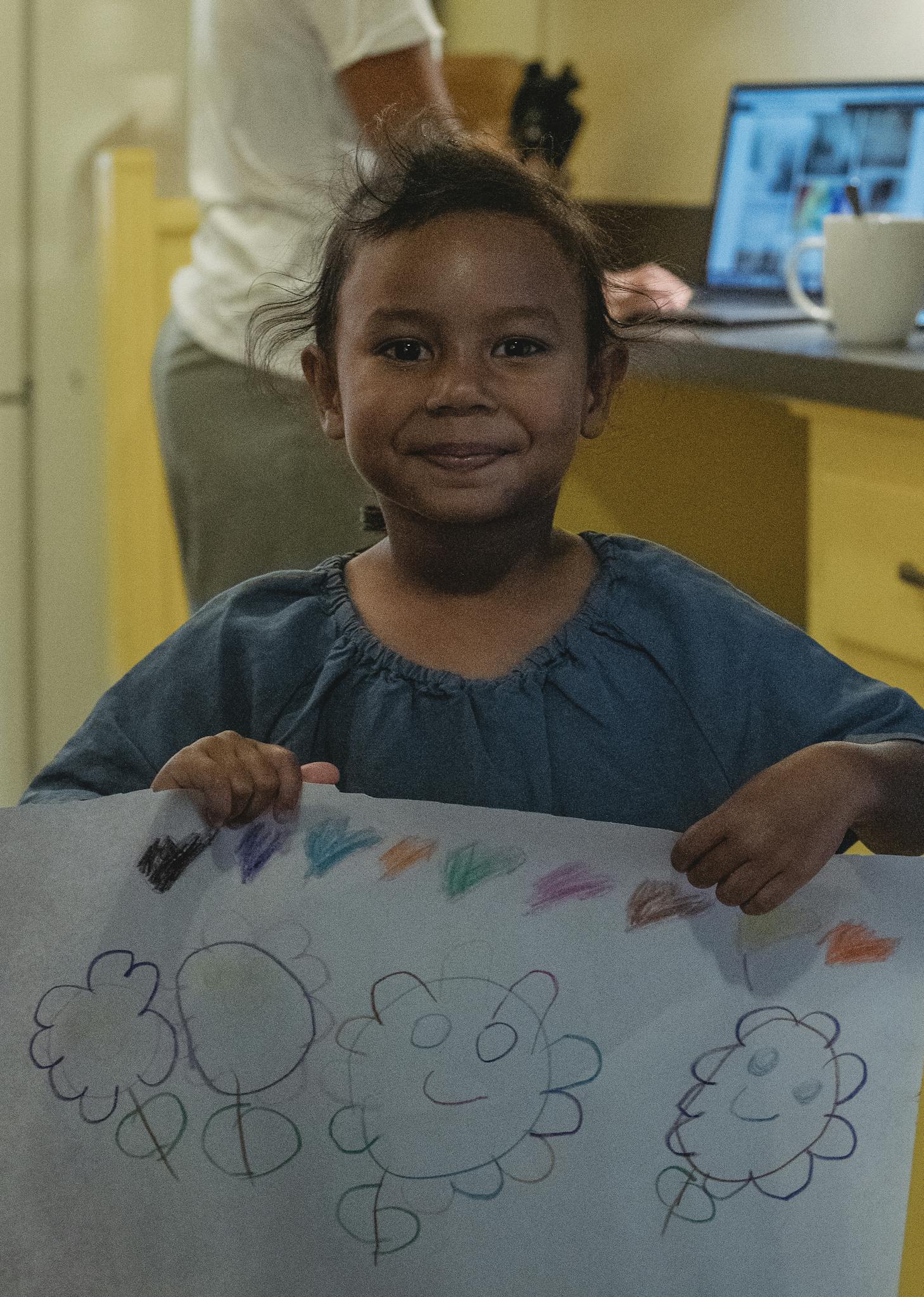 Smiling child proudly holding her colorful drawing in the kitchen setting.